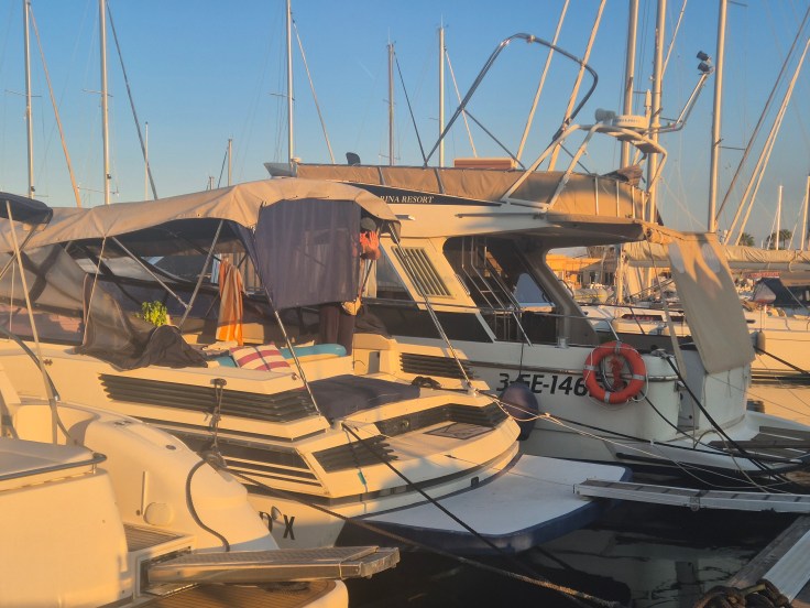 Two boats docked at a marina during sunset, with masts of other boats in the background.