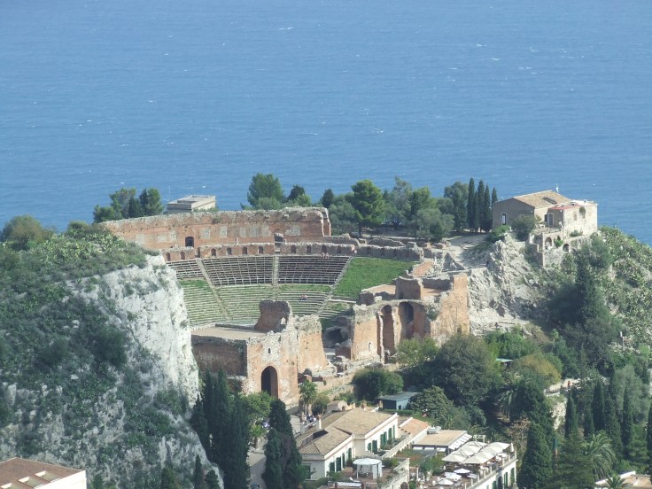 Aerial view of an ancient Roman theater ruins surrounded by greenery, with the sea in the background.