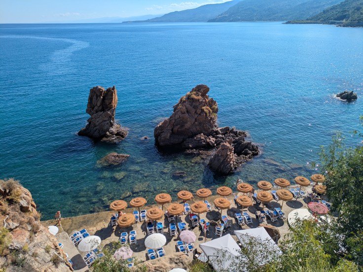 A scenic beach view featuring rocky formations in the water, sun loungers with umbrellas arranged on the shore, and calm blue waters, under a clear sky.