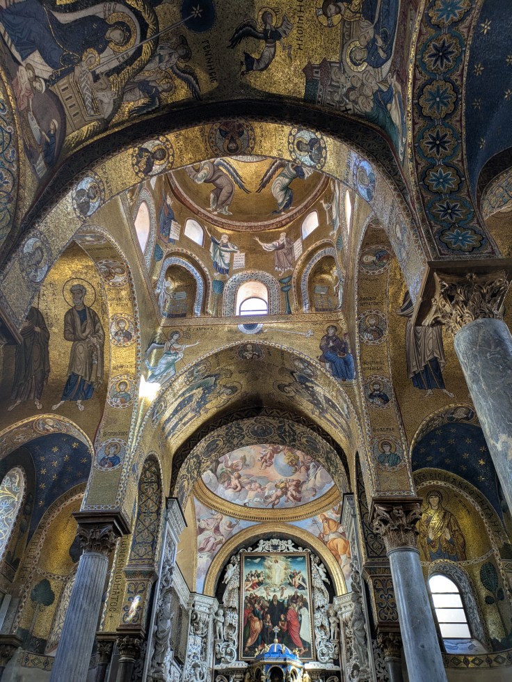 Interior view of a historic church with ornate golden mosaics and frescoes depicting religious figures and scenes on the vaulted ceilings and walls.