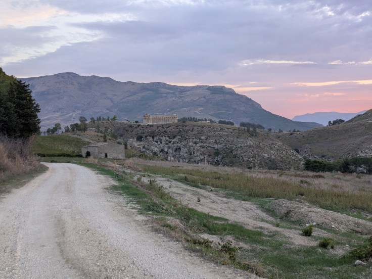 A gravel road winding through a landscape with mountains in the background, featuring a historic building on a hillside under a cloudy sunset sky.